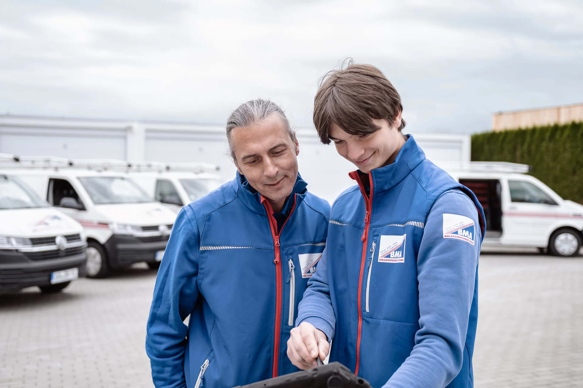 2 Mitarbeiter schauen sich am iPad gemeinsam den nächsten Auftrag an. Im Hintergrund ist die Fahrzeugflotte zu sehen.